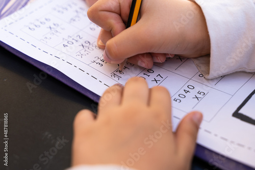 Close up on little girl hands doing school homework. Student writing with pencil maths multiplication exercise sheet