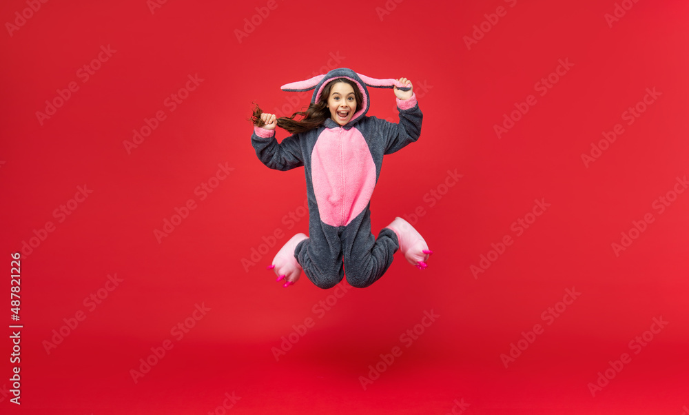 full length of amazed child in cozy sleepwear jump on red background, freedom