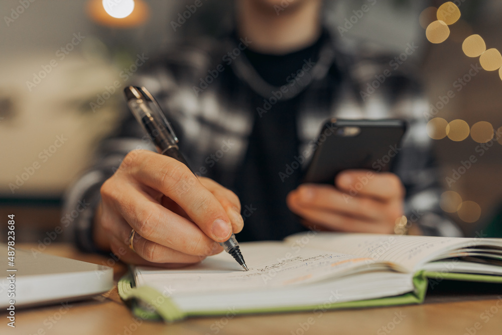 Crop male freelancer taking notes in notebook and browsing smartphone while working online in cafe Crop male freelancer taking notes in notebook and browsing smartphone while working online in cafe