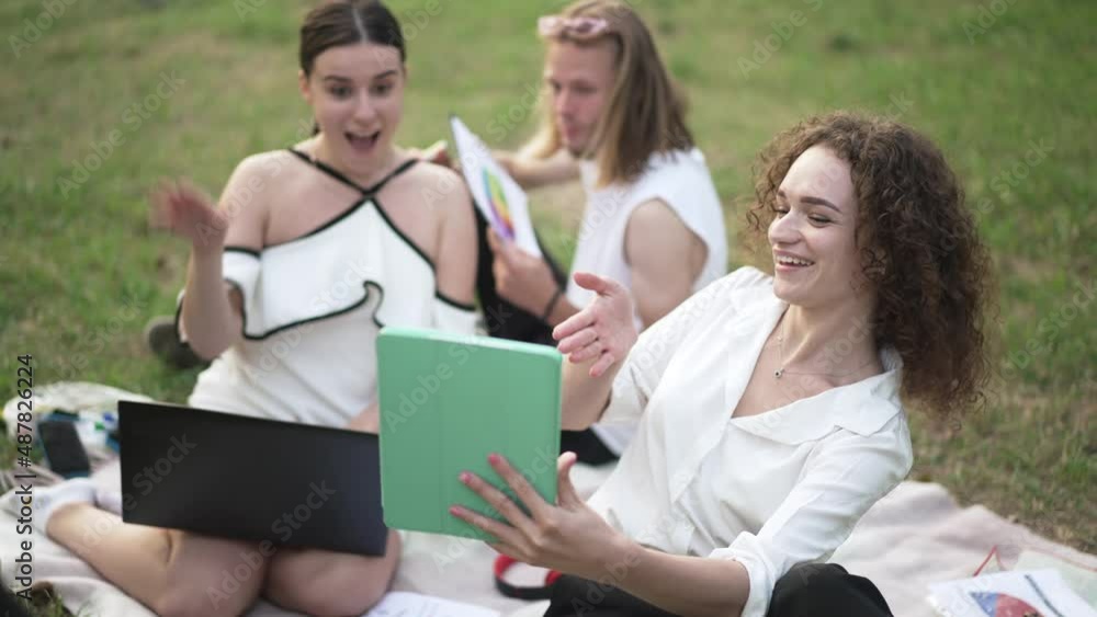 Team of startupers chatting online waving smiling talking at tablet video chat outdoors. Positive Caucasian woman bragging strategy planning online sitting on spring summer park meadow. Slow motion Team of startupers chatting online waving smiling talking at tablet video chat outdoors. Positive Caucasian woman bragging strategy planning online sitting on spring summer park meadow. Slow motion