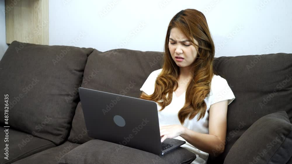 Young Asian woman sitting on sofa frowning and getting moody from working seriously on laptop in living room. Young Asian woman sitting on sofa frowning and getting moody from working seriously on laptop in living room.