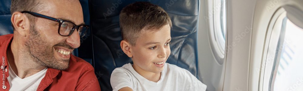 Happy little boy looking out the window, showing something to his father while sitting near the window, traveling by plane together. Family, transportation, vacation concept Happy little boy looking out the window, showing something to his father while sitting near the window, traveling by plane together. Family, transportation, vacation concept