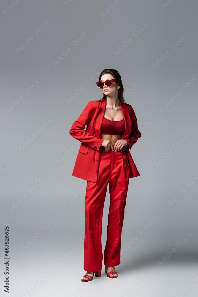 full length of young woman in trendy red suit and sunglasses posing on dark grey.