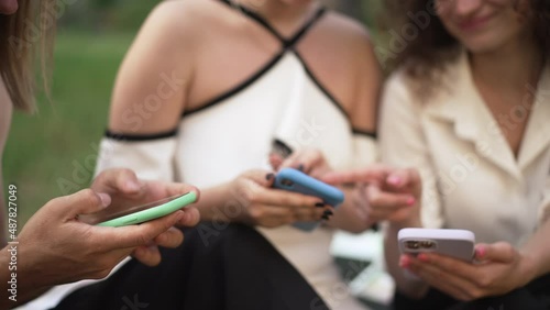 Three unrecognizable millennial man and women surfing Internet on smartphones talking sitting in sunny park. Young Caucasian friends gossiping meeting outdoors on weekend. Leisure and friendship