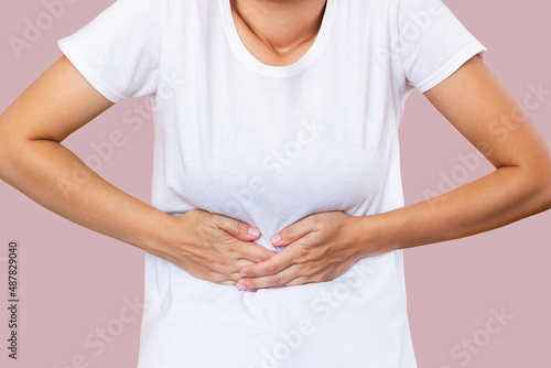 Cropped shot of a young woman in a white t-shirt holding her stomach with her hands isolated on color background. Stomach pain