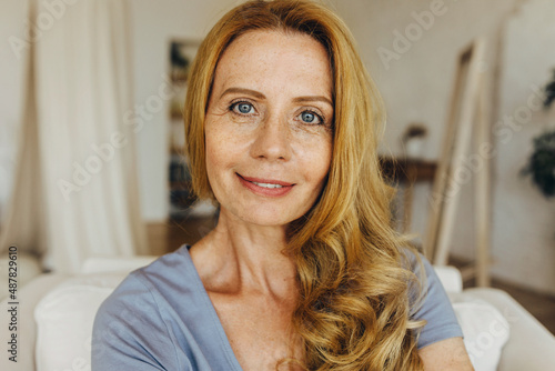 Caucasian adult woman of 50s wearing no makeup up, having natural beauty, perfect ageing face skin, covered with freckles, some face lines and wrinkles, sitting against home interior background