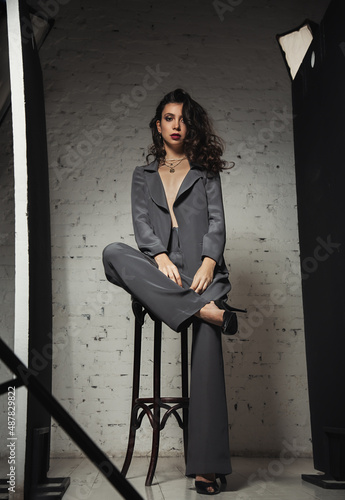 Sexy makeup beautiful model posing in fashion grey style suit clothing in high heels on the chair on brick decoration studio background.