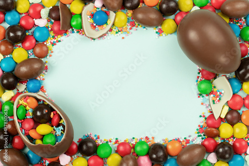 Frame of chocolate Easter eggs and colorful sweets on light blue background. Space for text.