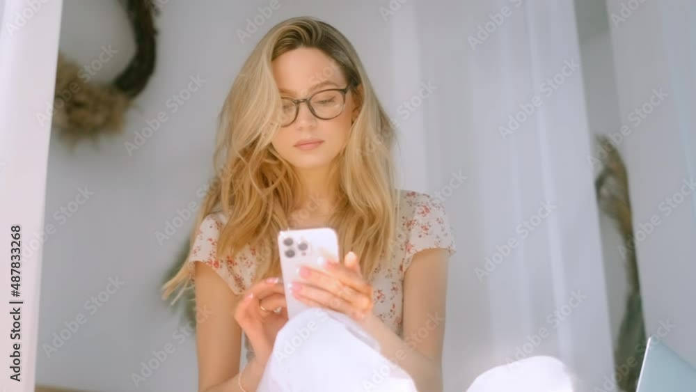 Happy smiling young attractive caucasian woman in eyeglasses using smartphone, sitting on a bed. Happy smiling young attractive caucasian woman in eyeglasses using smartphone, sitting on a bed.