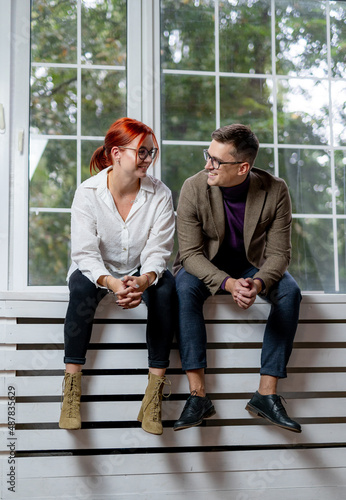 Man and woman sitting on the wooden platform. Big windows on the background. Red haired girl and brunette male.