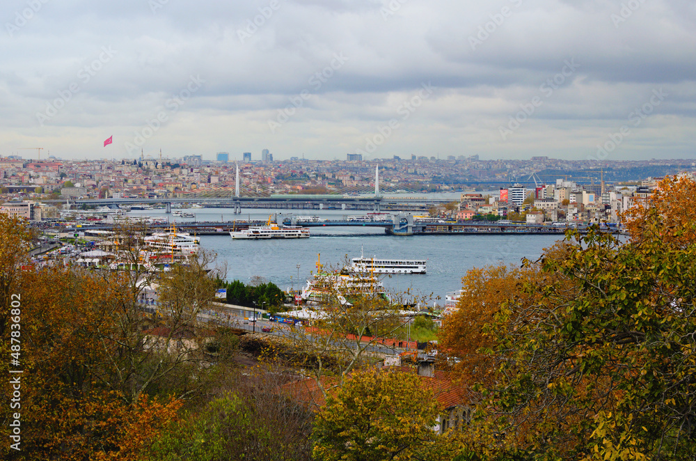 Istanbul, Turkey-October 25, 2021:Picturesque autumn landscape of The Golden Horn, Galata Bridge with many restaurants, Ataturk Bridge and buildings on horizon. Tree leaves border