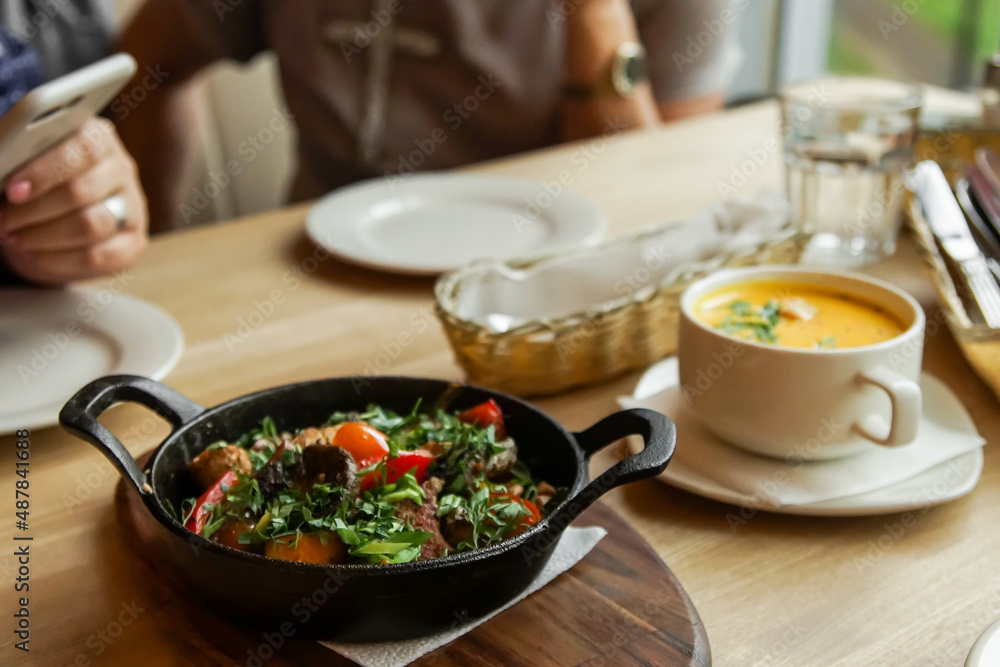 a frying pan with fried meat and vegetables in the foreground stands on a wooden table a frying pan with fried meat and vegetables in the foreground stands on a wooden table