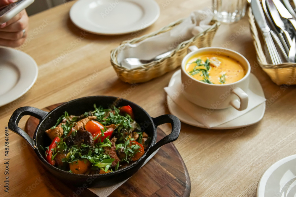a frying pan with fried meat and vegetables in the foreground stands on a wooden table a frying pan with fried meat and vegetables in the foreground stands on a wooden table