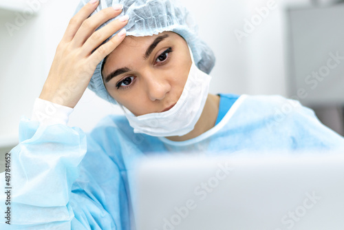 A tired female doctor wearing face mask and surgical overalls is sitting in front of a laptop screen in a medical office.Healthcare and medical concept.