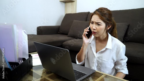Young Asian woman sitting on the floor and using laptop and use mobile phone for working in living room at home frowning and getting moody from working seriously.