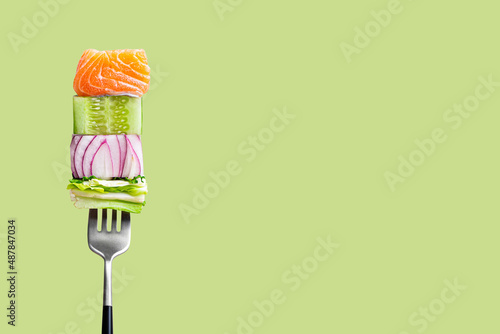 Close-up of fork with food on it: delicious fillet salmon, cucumber, onion, green salad on green background. Concept of healthy diet and clean eating, balanced nutrition space for text