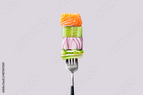 Close-up of fork with food on it: delicious fillet salmon, cucumber, onion, green salad on gray color background. Concept of healthy diet and clean eating with fish and vegetables, balanced nutrition
