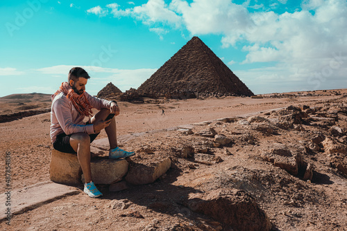 Latino man sitting on a rock in front of the giza pyramids, looking at his phone. Amazing sight of the great pyramids in cairo, egypt. Bright sunny winter day with blue sky