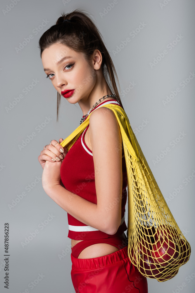 young stylish woman in red sportive outfit holding yellow string bag with apples isolated on grey. young stylish woman in red sportive outfit holding yellow string bag with apples isolated on grey.