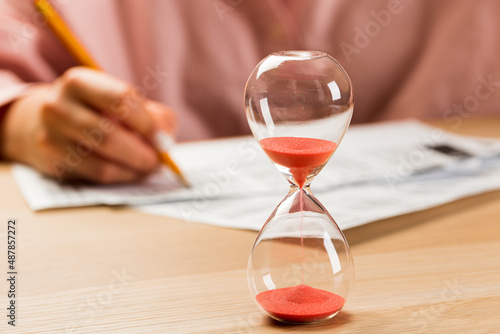 Hourglass with time running out and student hand testing in exercise and passing exam carbon paper computer sheet with pencil in school test room, education concept