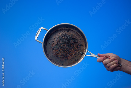 Dirty oily burnt metal frying pan or skillet held in hand by male hand. Close up studio shot, isolated on blue background photo