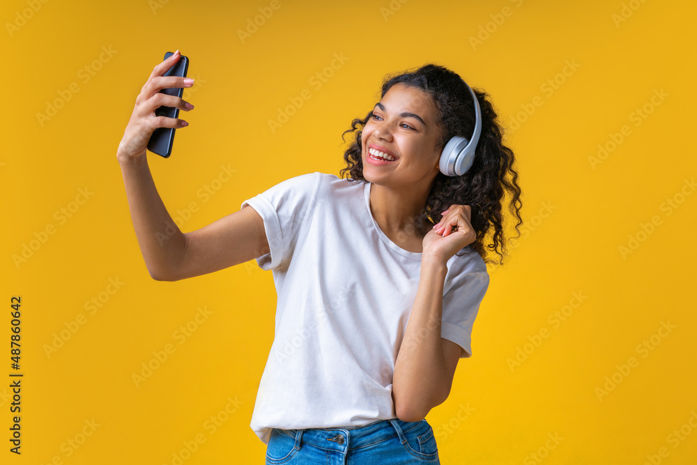 Studio portrait of happy smiling beautful brunette girl wearing wireless headphones and taking selfie on her mobile phone, isolated over yellow background Studio portrait of happy smiling beautful brunette girl wearing wireless headphones and taking selfie on her mobile phone, isolated over yellow background