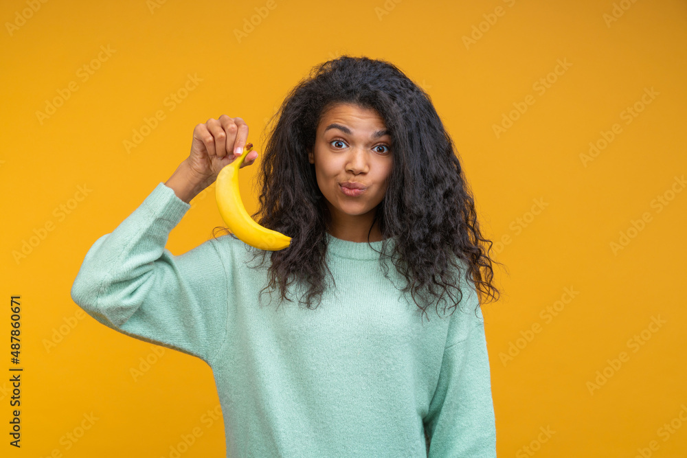 Studio portrait of cute and funny african american girl posing with a banana in hand, isolated over bright colored yellow backgorund