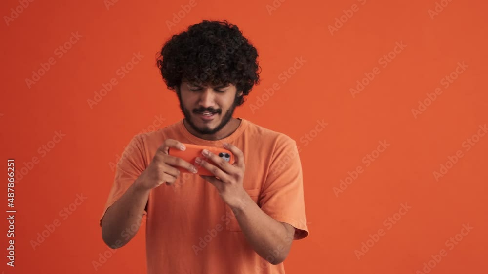 Concentrated curly-haired Indian man playing game by phone and rejoicing about victory in the orange studio Concentrated curly-haired Indian man playing game by phone and rejoicing about victory in the orange studio