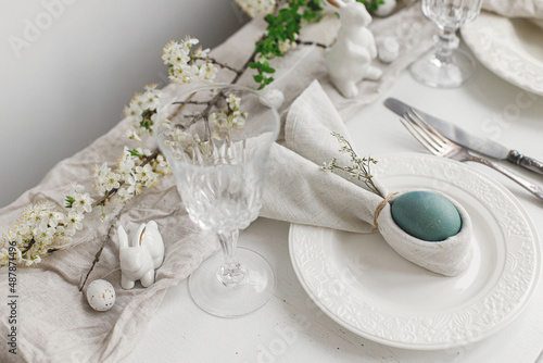 Stylish elegant Easter brunch table setting. Easter egg in bunny napkin on plate with cutlery, bunny, spring flowers on rustic white table. Modern Easter table arrangement