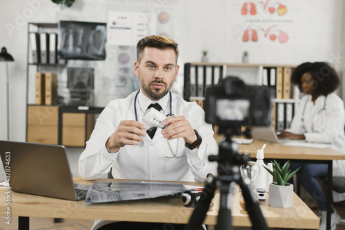Caucasian medical worker sitting at desk with electronic thermometer in hands and recording video on professional camera. Telemedicine, people and technology concept.