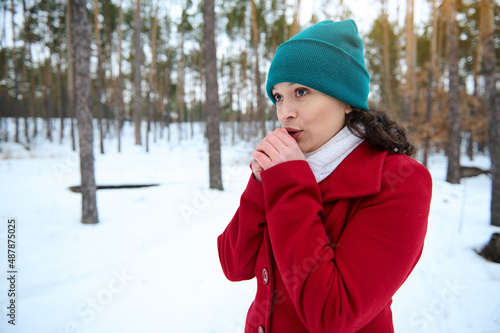 Charming young Caucasian woman in knit green hat and warm cozy woolen red coat holding her hands crossed because of cold weather and blowing on it for warming them while standing in the snowy forest