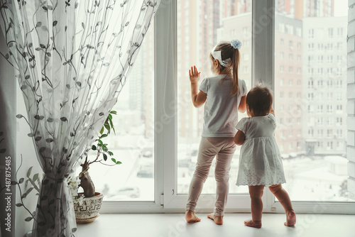 Two little girls, sisters are standing on the windowsill and looking out the window. Outside the window is a winter city covered with snow.