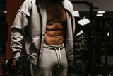 A bodybuilder in an opened hoodie is showing his abs while holding dumbbells.