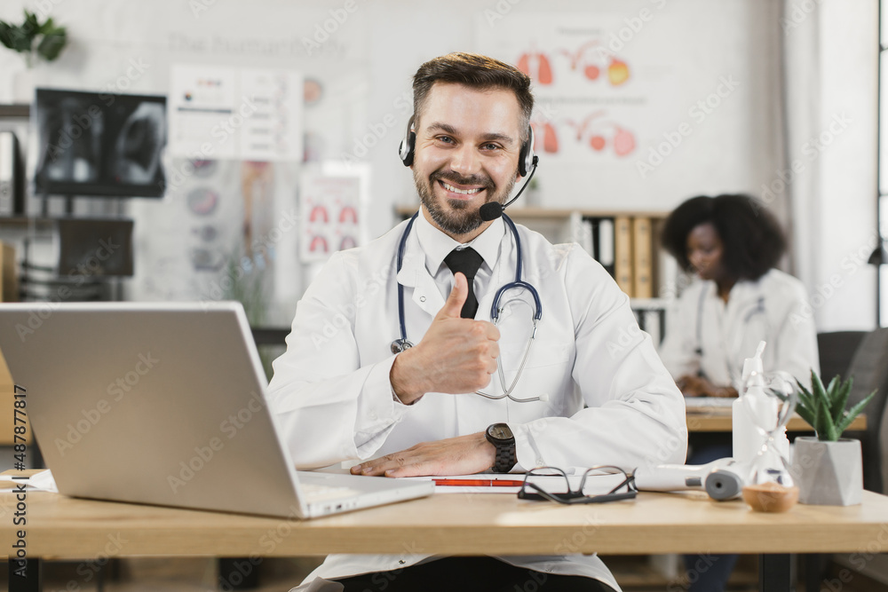 Competent caucasian doctor in headset and lab coat sitting at desk and looking at camera during video chat on modern laptop and showing thumb up. African american nurse sitting on background. Competent caucasian doctor in headset and lab coat sitting at desk and looking at camera during video chat on modern laptop and showing thumb up. African american nurse sitting on background.