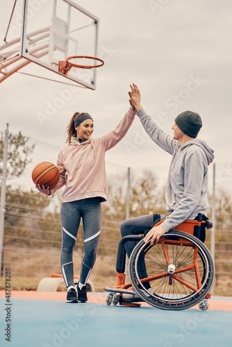 Wheelchair bound athlete and his female friend giving high five after playing basketball outdoors.