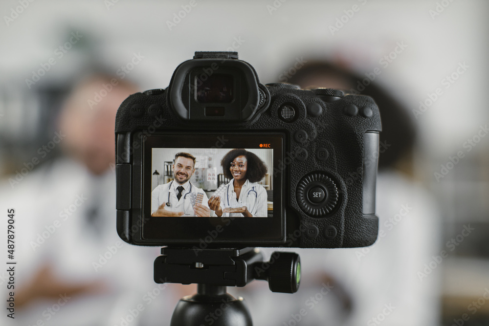 Two multiracial doctors in white lab coats holding packs of pills while recording video blog on professional camera. Focus on screen, blur background. Two multiracial doctors in white lab coats holding packs of pills while recording video blog on professional camera. Focus on screen, blur background.