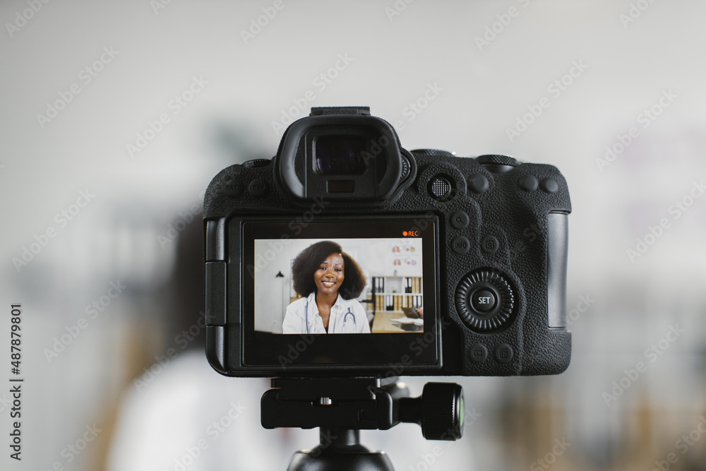 Video camera screen with smiling african american female doctor in white lab coat sitting a hospital office. Competent medical worker creating content for her channel.