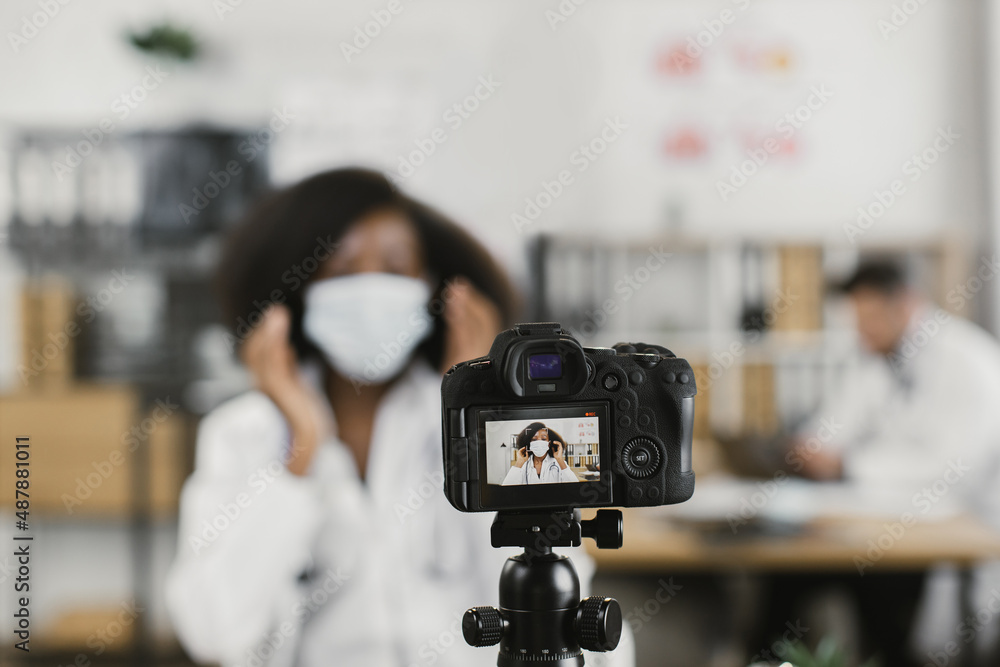 African american woman in lab coat filming online tutorial about proper wearing of medical face mask. Blur background of caucasian male assistant working on laptop.