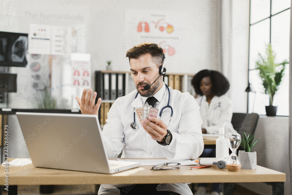 Caucasian male doctor in headset having online meeting with colleagues about clinical trials of the drug. Medical specialist sitting at desk with various pills in hands and looking on laptop screen.