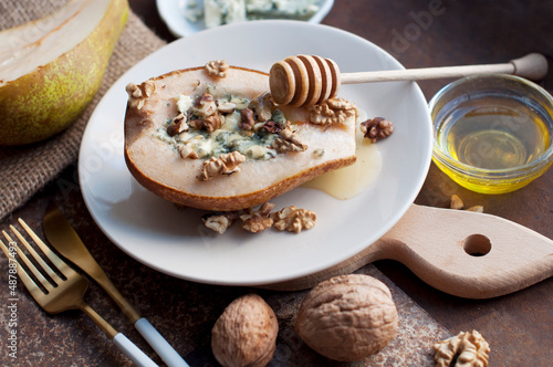 Pear baked with blue cheese, honey and walnuts on a plate on a dark brown background. Backed pear with fork, knife and wooden honey stick. Rustic style
