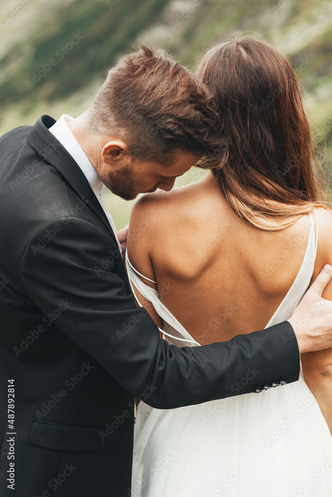 Close-up portrait of a groom kissing the shoulder of his bride standing with her back at camera wearing a dress with her back uncovered. Close-up portrait of a groom kissing the shoulder of his bride standing with her back at camera wearing a dress with her back uncovered.