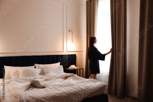 Beautiful young woman wearing silk robe near window in hotel room