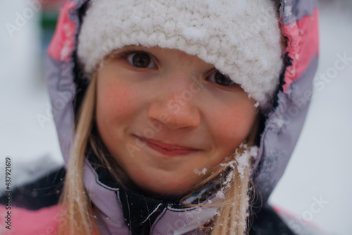 Close-up portrait of happy blond pretty preschool girl wearing bright colored snowsuit and white hat in a cold winter day. Selective focus