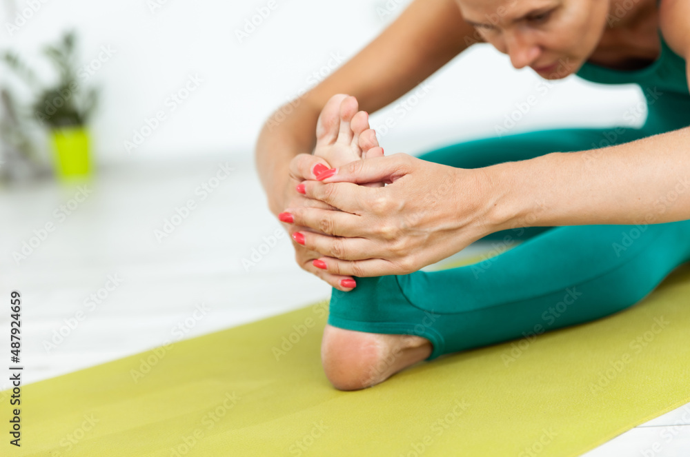 Yogi woman in sports clothes practices leg stretching, holding the heel with her hand while sitting on the mat in yoga studio