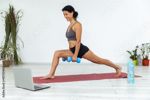 Online fitness class. Young athletic woman trainer exercising with dumbbell, broadcasting with laptop, recording her fitness blog or streaming indoors