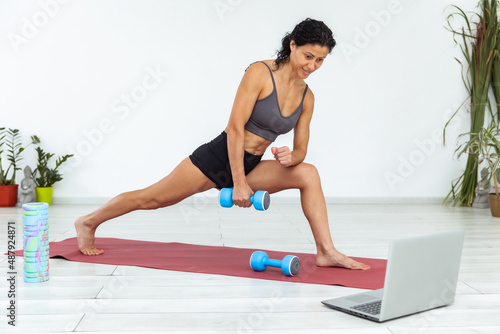 Online fitness class. Young athletic woman trainer exercising with dumbbell, broadcasting with laptop, recording her fitness blog or streaming indoors