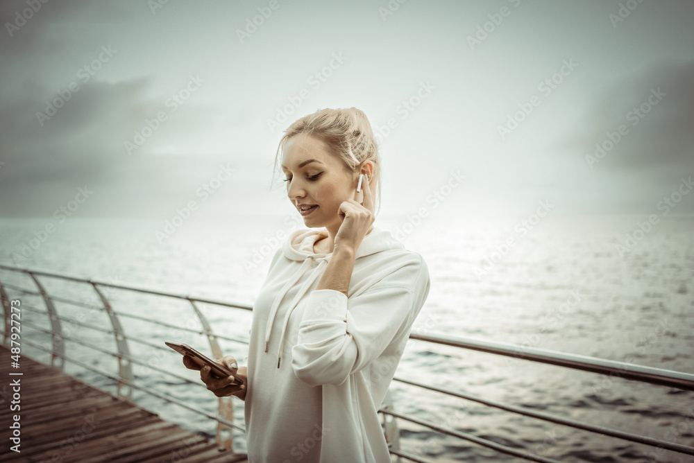 Young fitness woman blonde in a white hoodie uses a smartphone and listens to music on headphones on the beach Young fitness woman blonde in a white hoodie uses a smartphone and listens to music on headphones on the beach