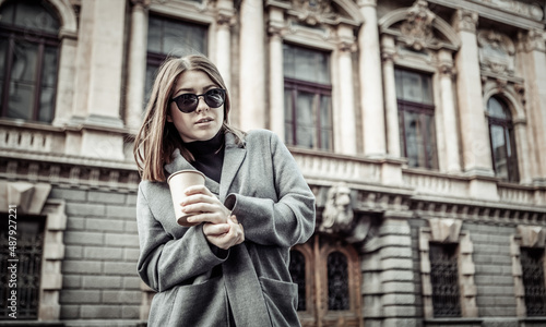 Dramatic portrait of a fashionable woman in an autumn coat and sunglasses. Attractive girl holding cup of coffee on the background of urban architecture