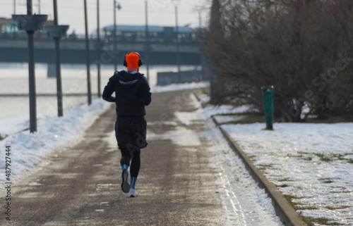 Defocused runner with bright orange hat and headphones run next to river in city center in morning