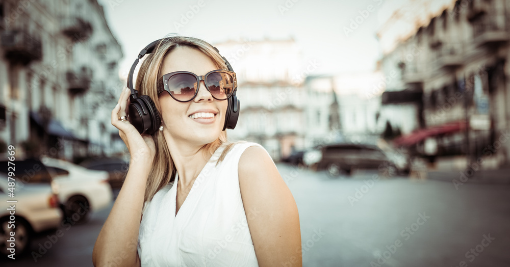 Young cheerful woman in sunglasses listening to music with headphones in the city